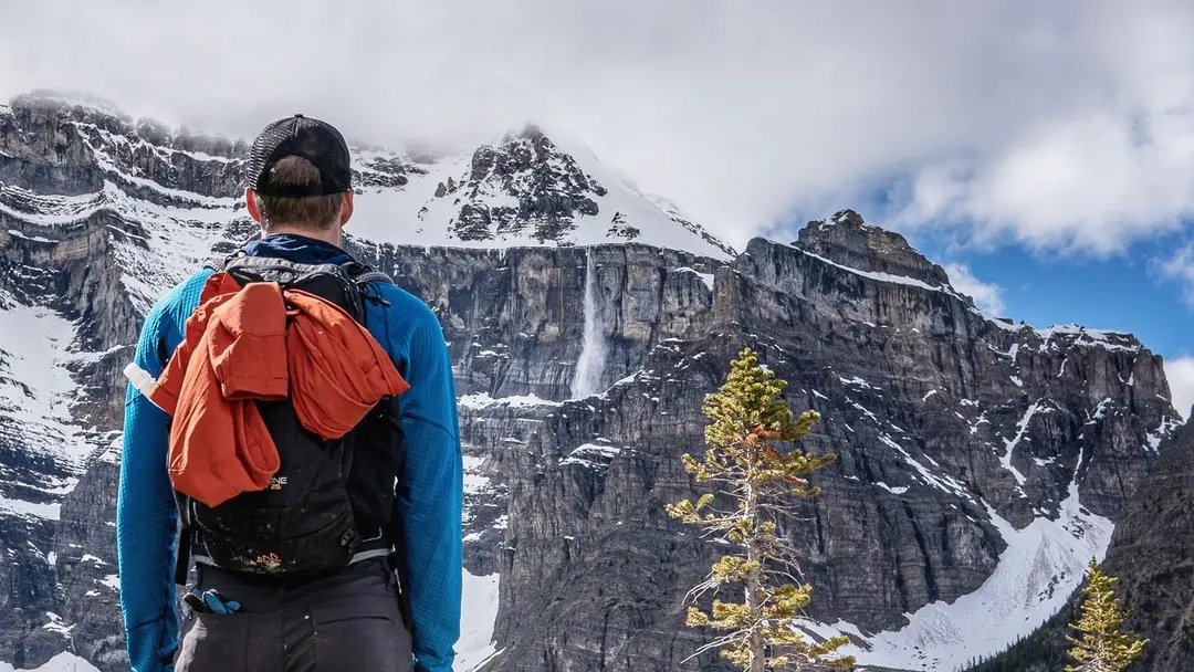 A hiker watches an avalanche release on a snow-loaded cliff face.