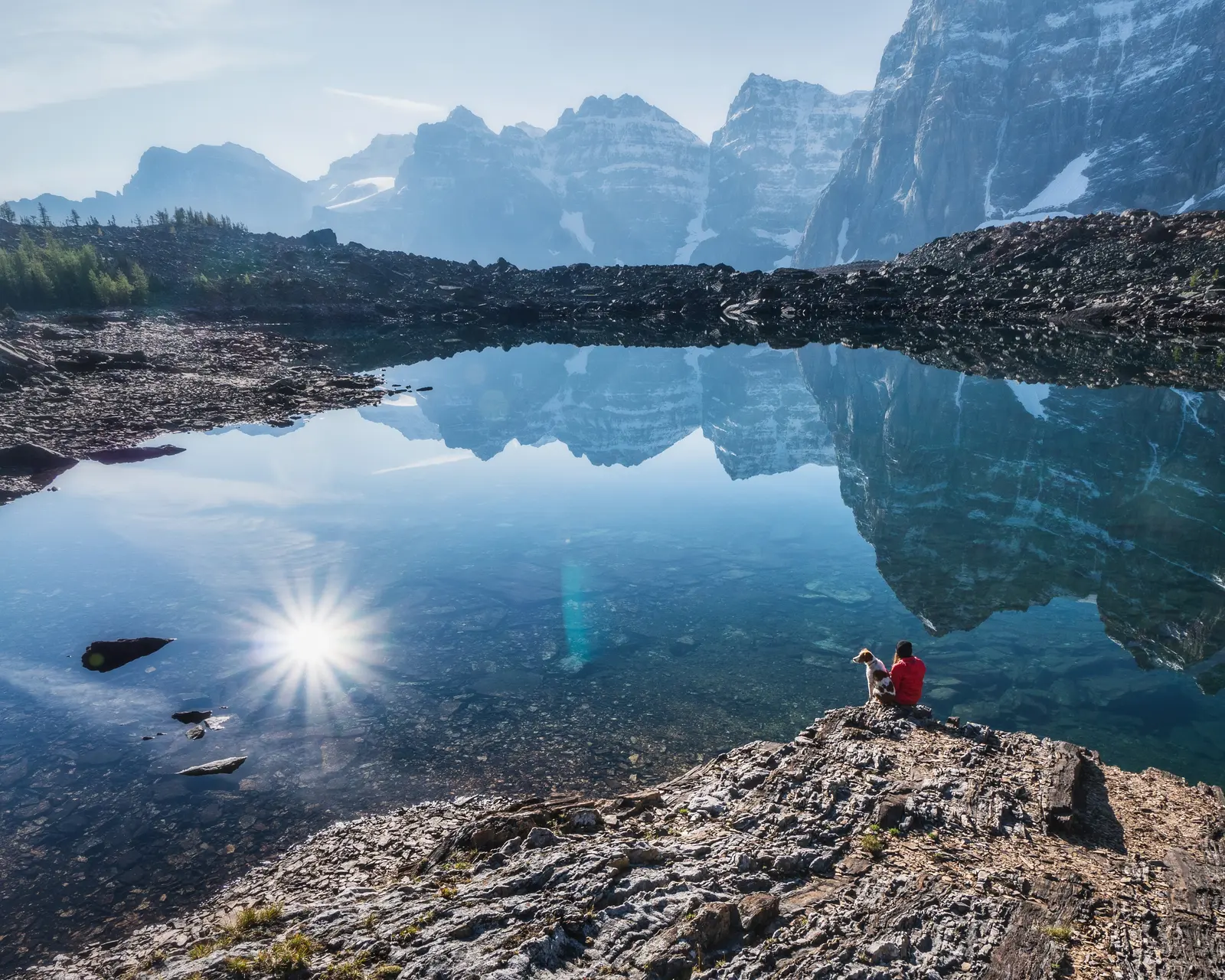 A hiker sitting with a dog beside Eiffel Lake below steep mountain slopes.