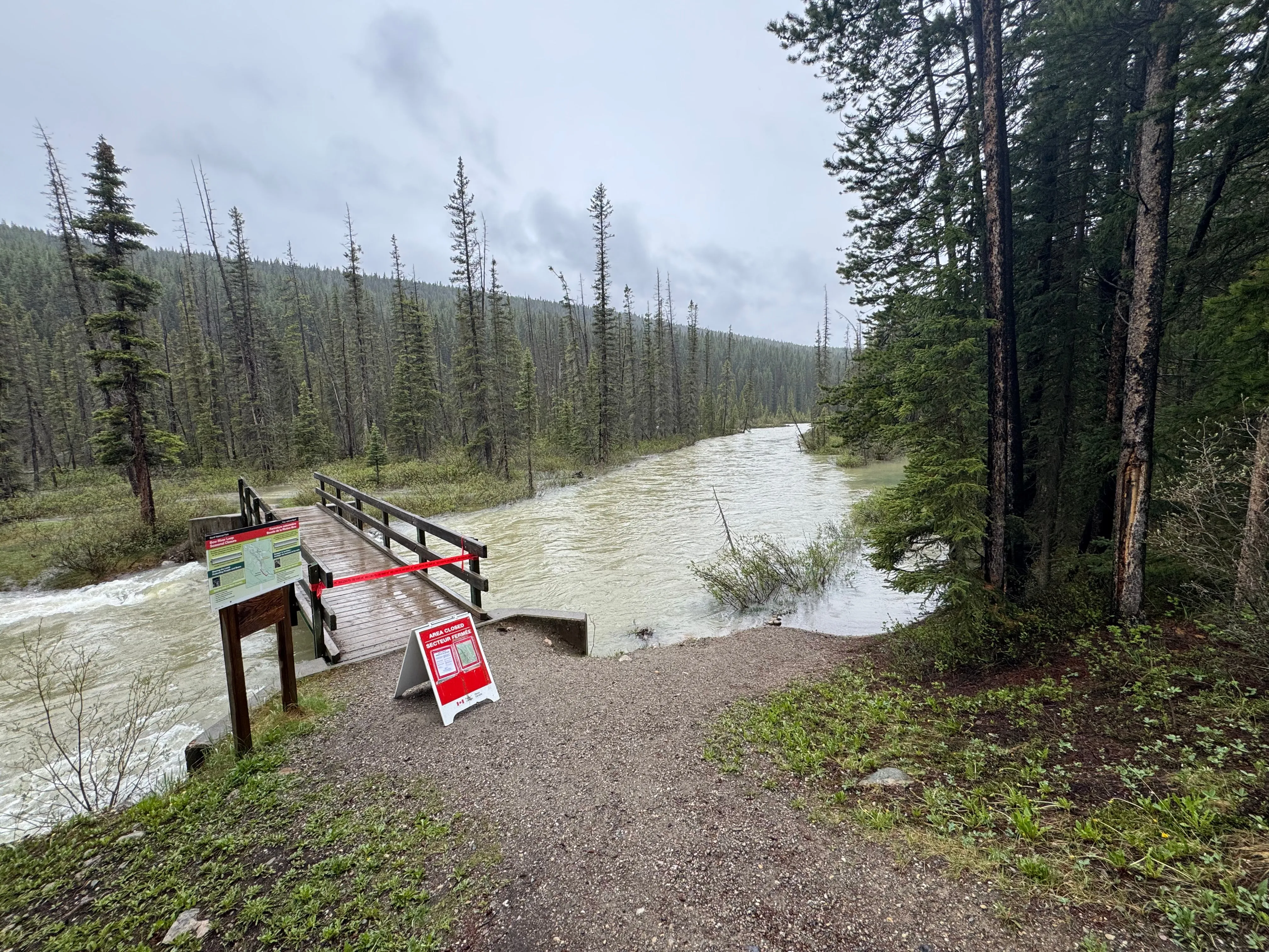 Closed pedestrian bridge over a swollen river with Parks Canada closure signs and red tape.