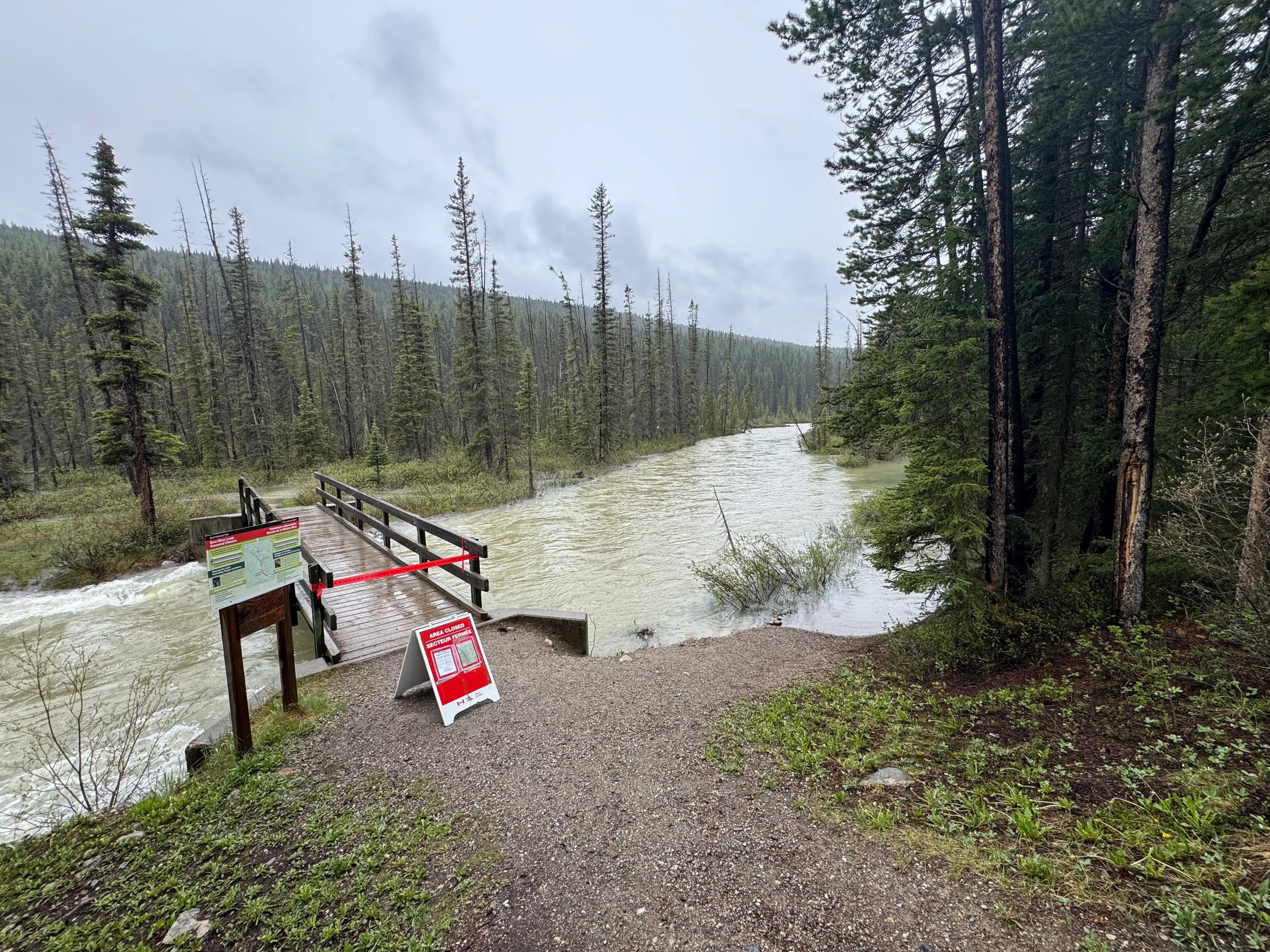 Closed pedestrian bridge over a swollen river with Parks Canada closure signs and red tape.