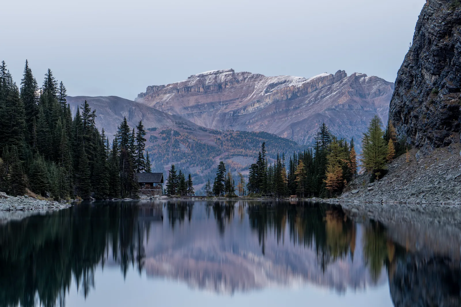 Lake Agnes with calm water reflecting the surrounding forest and mountains at dusk.