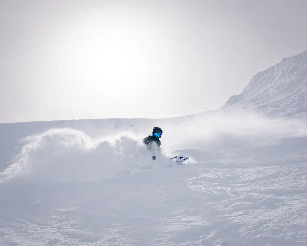A skier dressed in black with blue mirrored goggles carving through deep powder, throwing up a wide wave of snow, with a rounded ridge and an orange trail marker behind under flat, bright light.